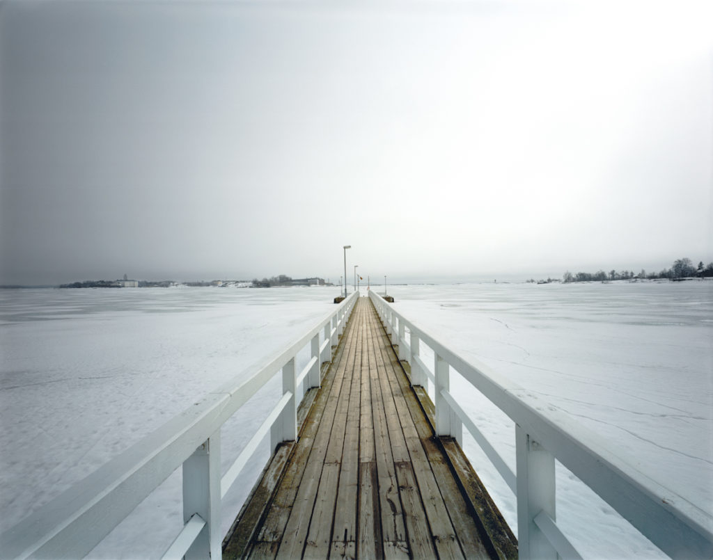 Imagen de un muelle de madera que se extiende sobre un lago congelado en un paisaje invernal, capturada con equipos profesionales para fotografía y vídeo, mostrando un cielo nublado y un entorno nevado.
