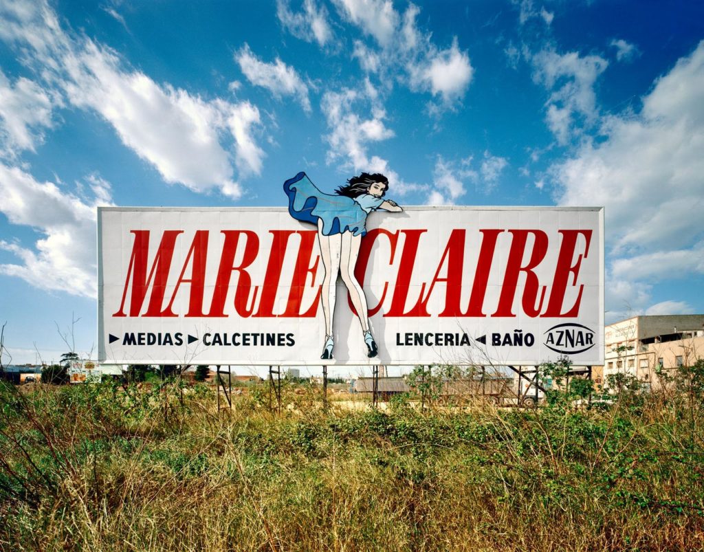 Imagen de una valla publicitaria de Marie Claire, especializada en medias, calcetines, lencería y baño, ubicada en un campo abierto con un cielo azul y nubes esponjosas, capturada con equipos profesionales para fotografía y vídeo.