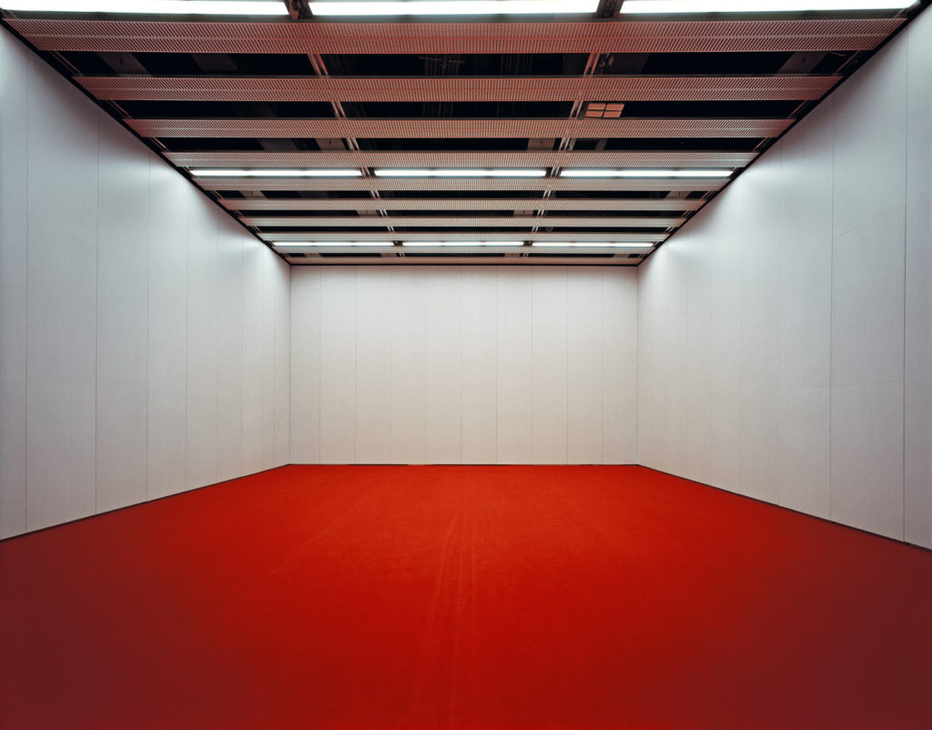 Imagen de una sala vacía con una alfombra roja y paredes blancas, capturada con equipos profesionales para fotografía y vídeo, mostrando un techo con iluminación fluorescente.