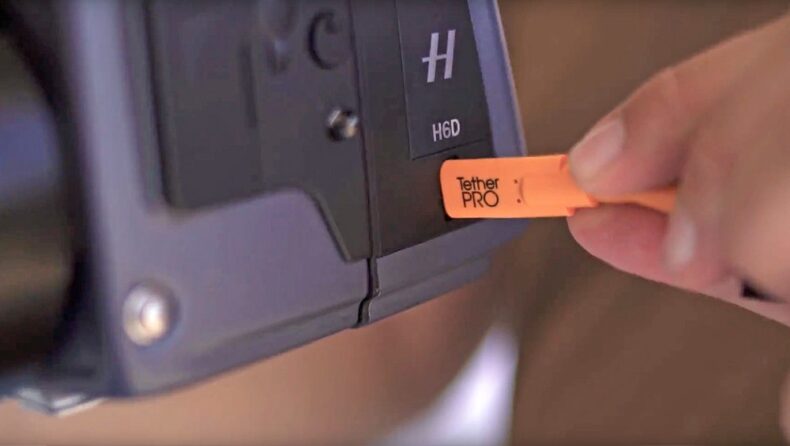 Mano insertando un cable TetherPro USB naranja en la cámara profesional Hasselblad H6D, destacando la importancia de los accesorios de conexión en equipos de fotografía y vídeo profesional.