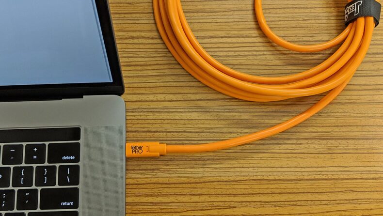 Cable Tether Pro naranja conectado a un portátil para transferencia de datos durante sesiones profesionales de fotografía y vídeo, sobre una mesa de madera.