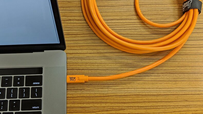 Cable TetherPro USB naranja conectado a un portátil, facilitando la transferencia de datos desde una cámara profesional, mostrando la integración de equipos de fotografía y vídeo en un entorno de trabajo digital.