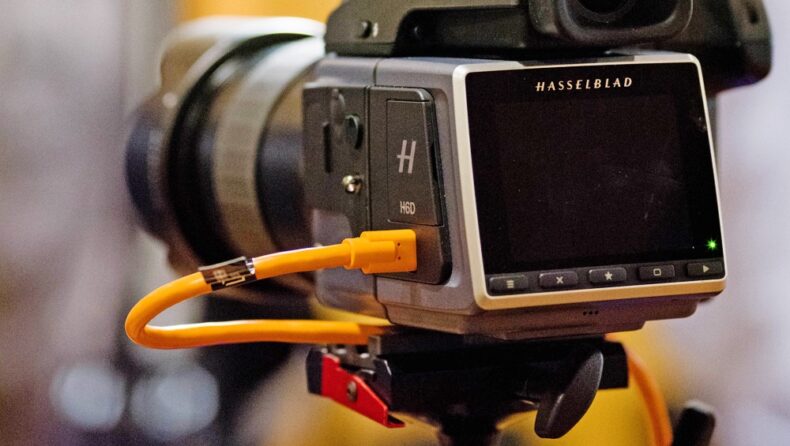Cámara Hasselblad H6D profesional conectada a través de un cable de tethering naranja para transmisión de datos rápida y segura, mostrando la pantalla trasera y el lente en un entorno de estudio fotográfico.