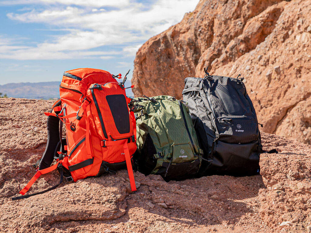 Tres mochilas de la serie montaña de f-stop en colores naranja, verde y negro, ubicadas sobre una formación rocosa en un entorno montañoso, ideales para fotógrafos aventureros y amantes del outdoor.