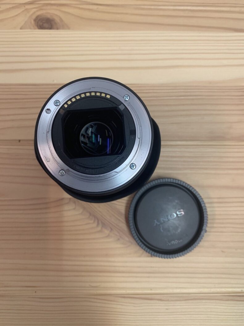 Vista trasera de un lente Carl Zeiss Sonnar FE 55mm f/1.8, mostrando los contactos electrónicos y con la tapa trasera Sony al lado sobre una mesa de madera, ideal para equipos profesionales de fotografía y vídeo.