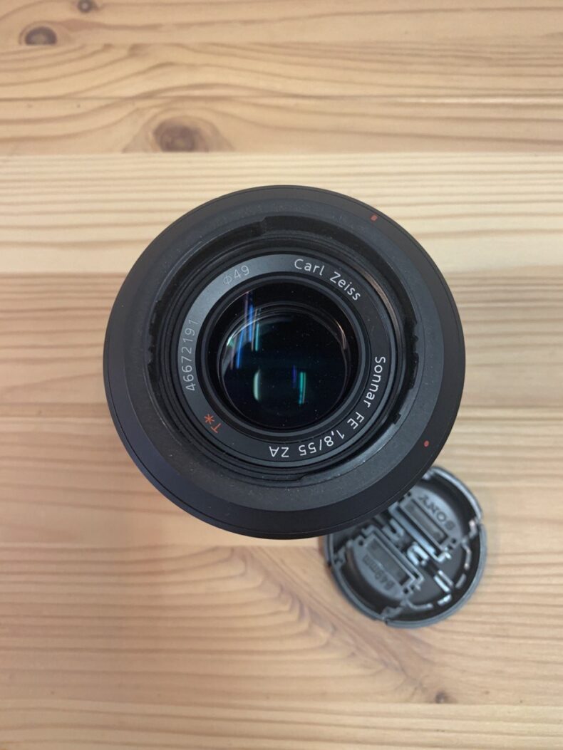 Vista superior de un lente Carl Zeiss Sonnar FE 55mm f/1.8, ideal para equipos profesionales de fotografía y vídeo, con la tapa del objetivo al lado sobre una mesa de madera.